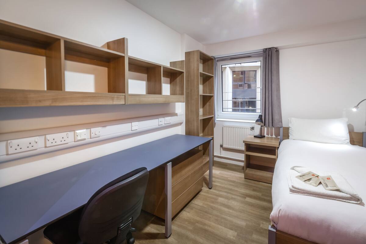 A modern hotel room with a wooden desk, a computer, a chair, a bed with a white comforter, a nightstand, and a window with curtains.