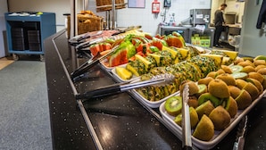 Buffet com diversas frutas e legumes, um relógio na parede e uma pessoa ao fundo.