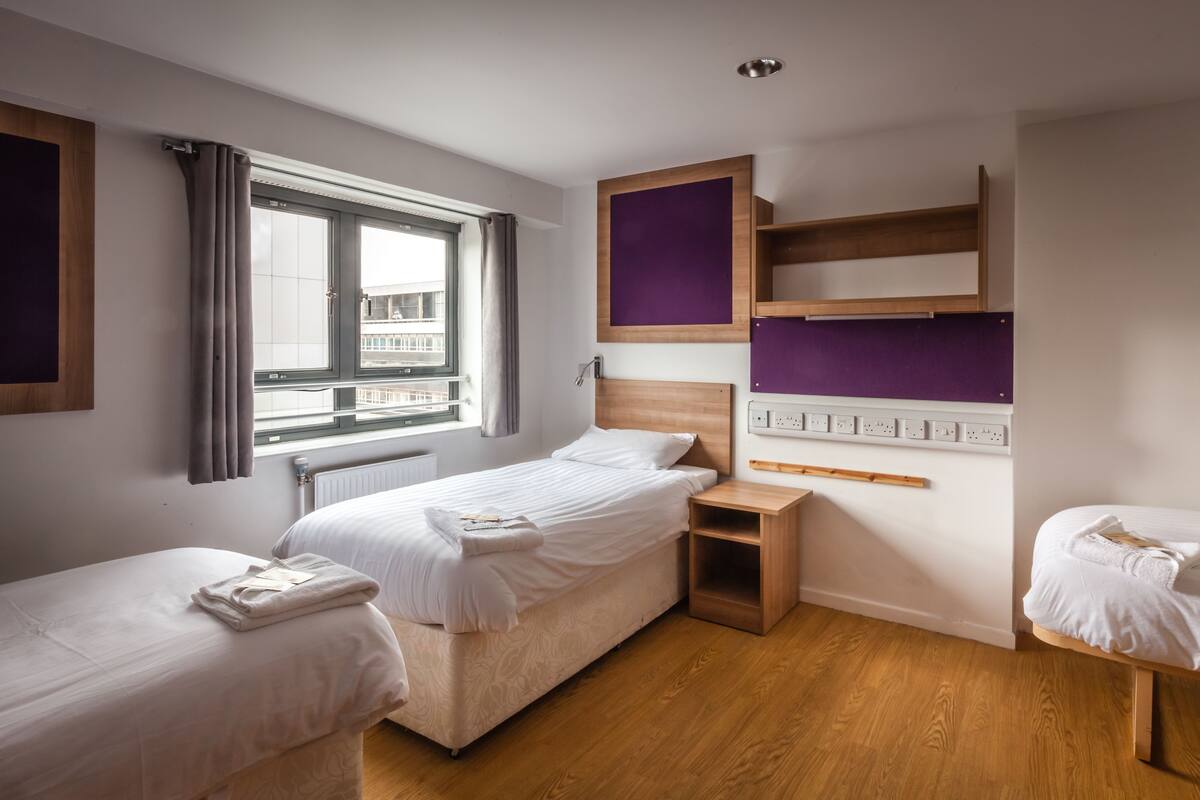 A hotel room with two beds, a window with curtains, a wooden headboard, a purple accent wall, and a wooden shelf.