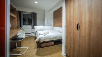 A modern bedroom with a wooden wardrobe, a bed with white linens, a desk with a chair, and a window with curtains.