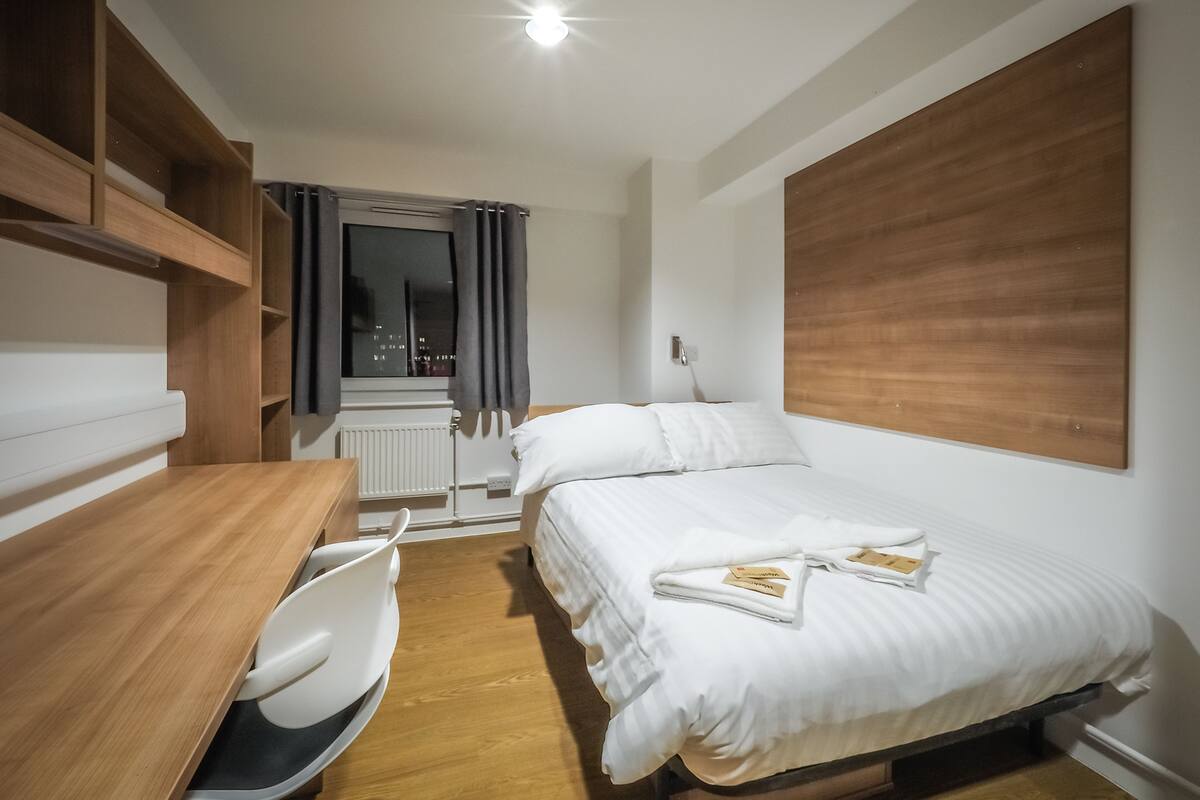 A modern hotel room with a wooden desk, a bed with white linens, a large window with curtains, and a view of the city at night.