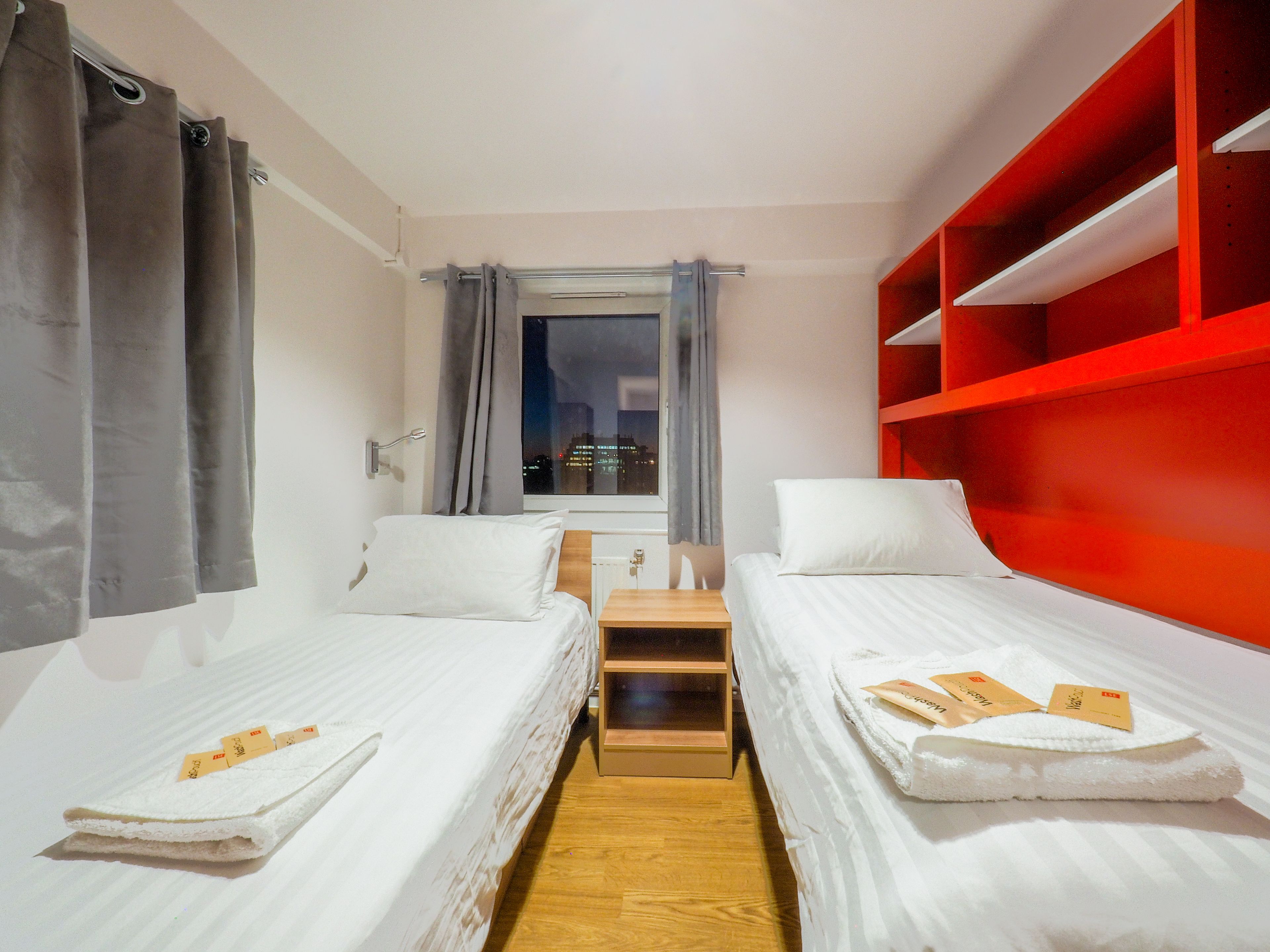 A hotel room with two single beds, a wooden nightstand, a window with curtains, and a red shelving unit.