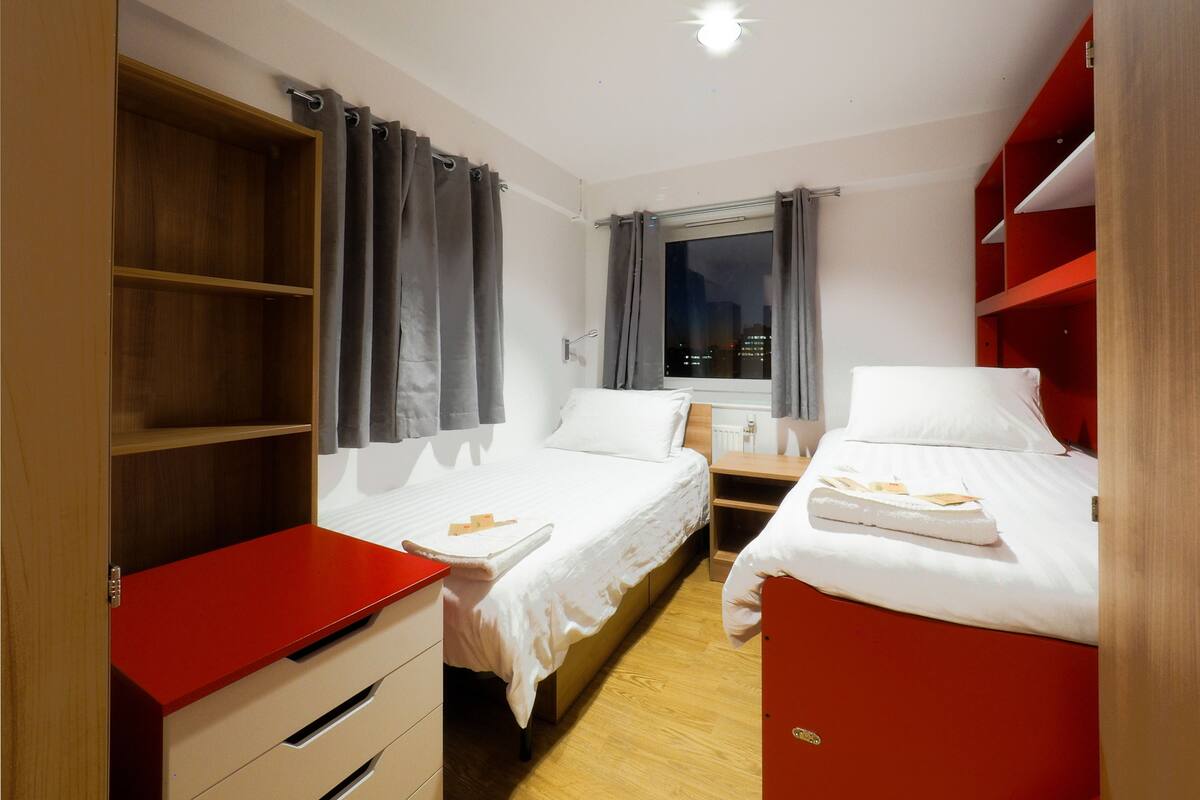 A small, well-lit room with two beds, a window with curtains, and a red storage unit.