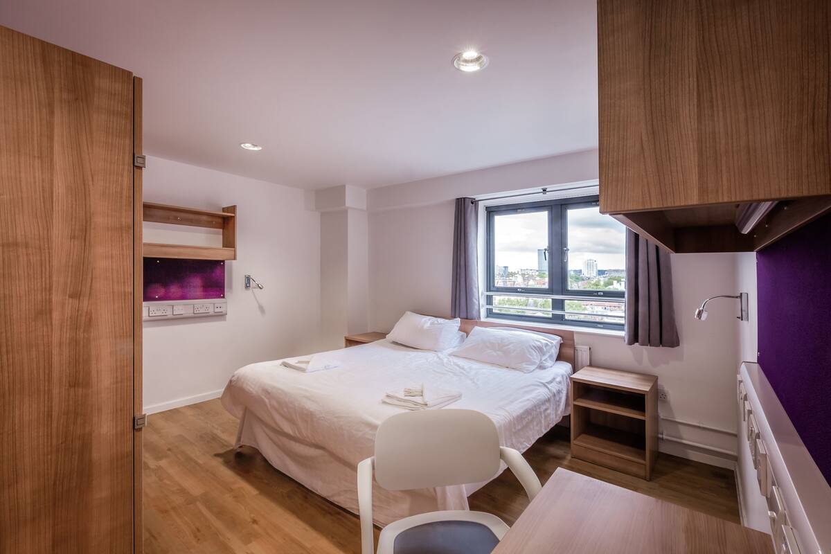 A modern hotel room with a bed, a desk, a chair, a window with a city view, and a built-in shelf.