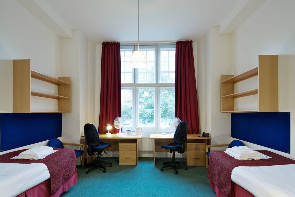 A room with two beds, a desk, two chairs, a computer, and a window with curtains.