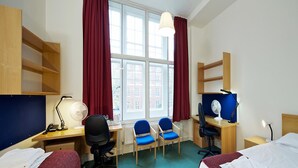 Habitación de hotel con dos camas, un escritorio con computadora, una silla, lámpara de escritorio, ventilador y ventana con cortinas.