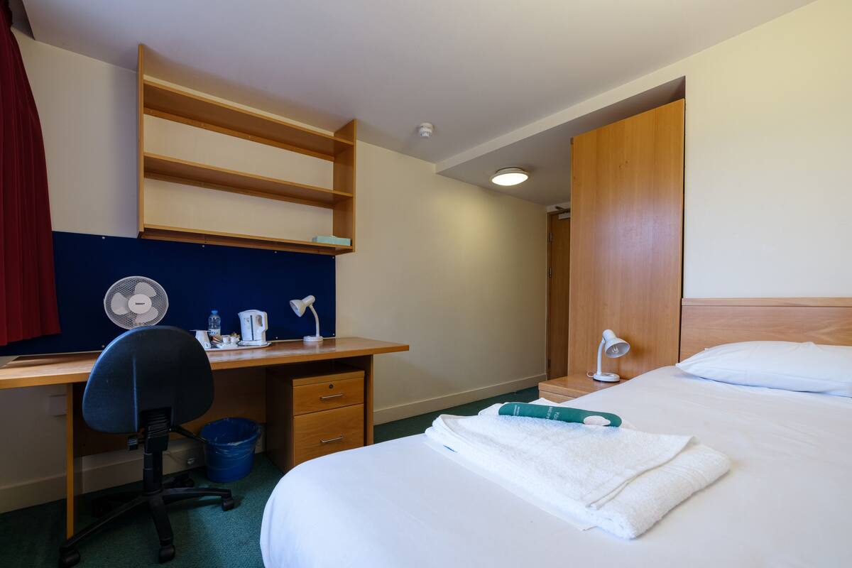 A hotel room with a bed, a desk with a chair, a desk lamp, a fan, and a wooden wardrobe.