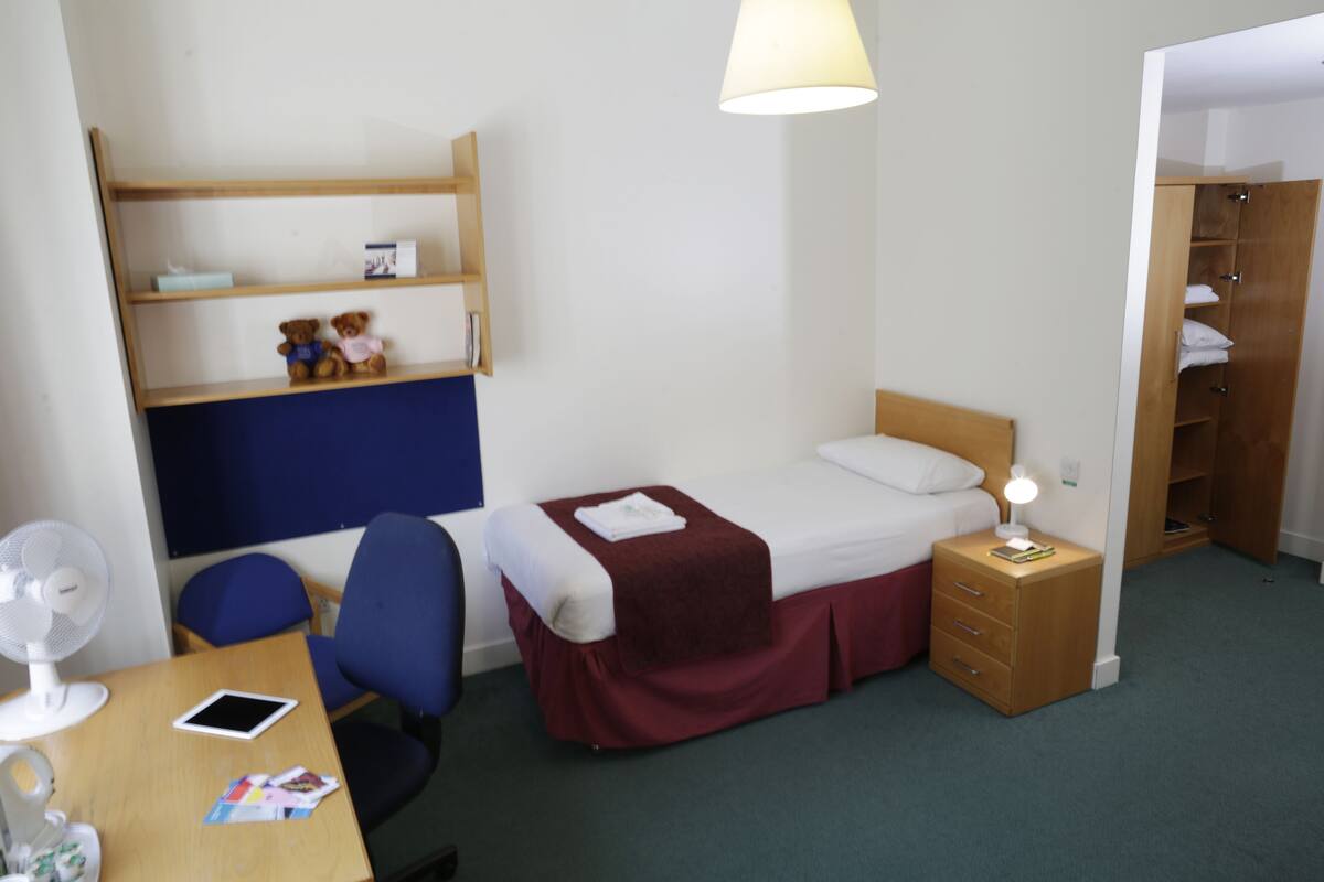 A small, neatly arranged room with a bed, bedside table, a desk with a chair, a shelf with teddy bears, and a wardrobe.
