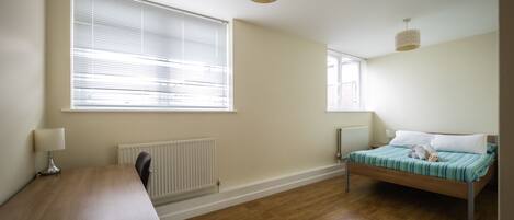 A bedroom with a wooden floor, a bed with a striped bedspread, a desk with a lamp, and two windows with blinds.