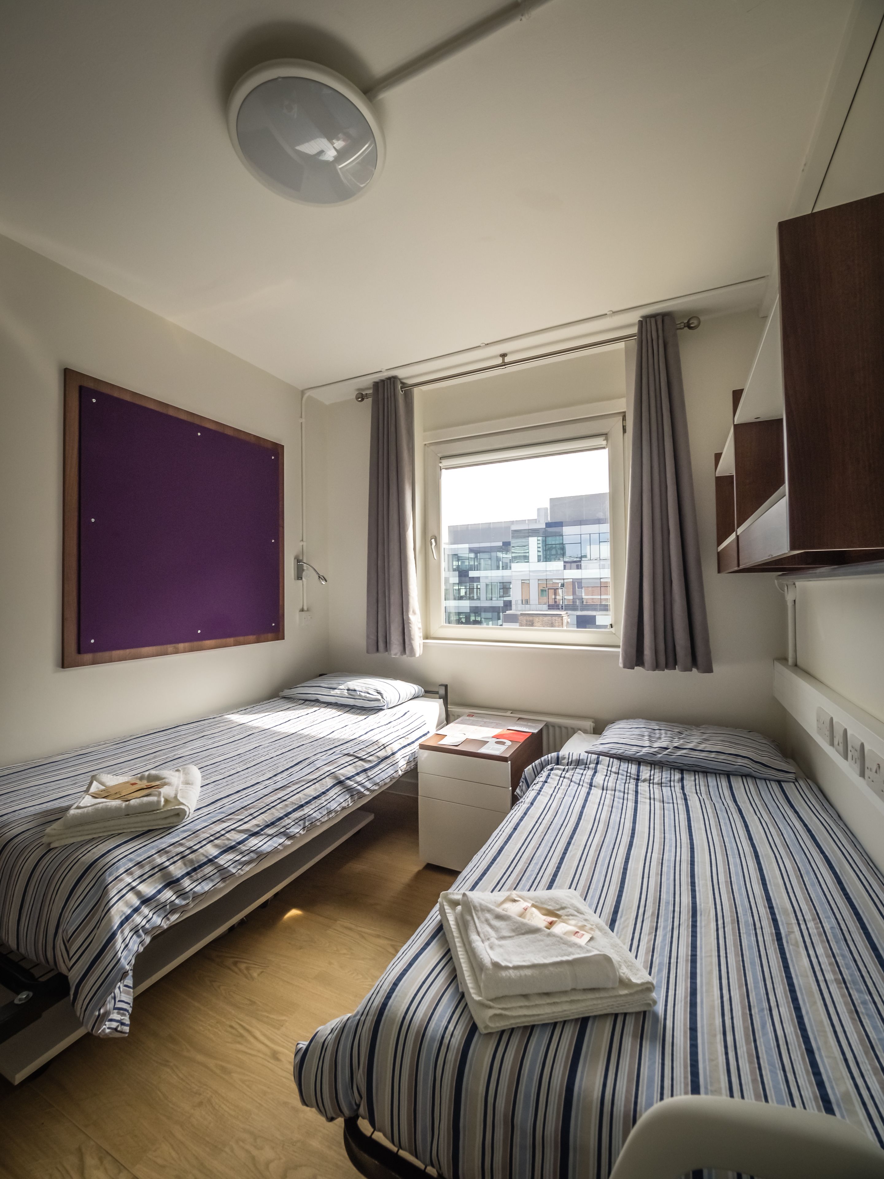 A hotel room with two single beds, a small desk, a window with curtains, a ceiling light, and a view of buildings outside.