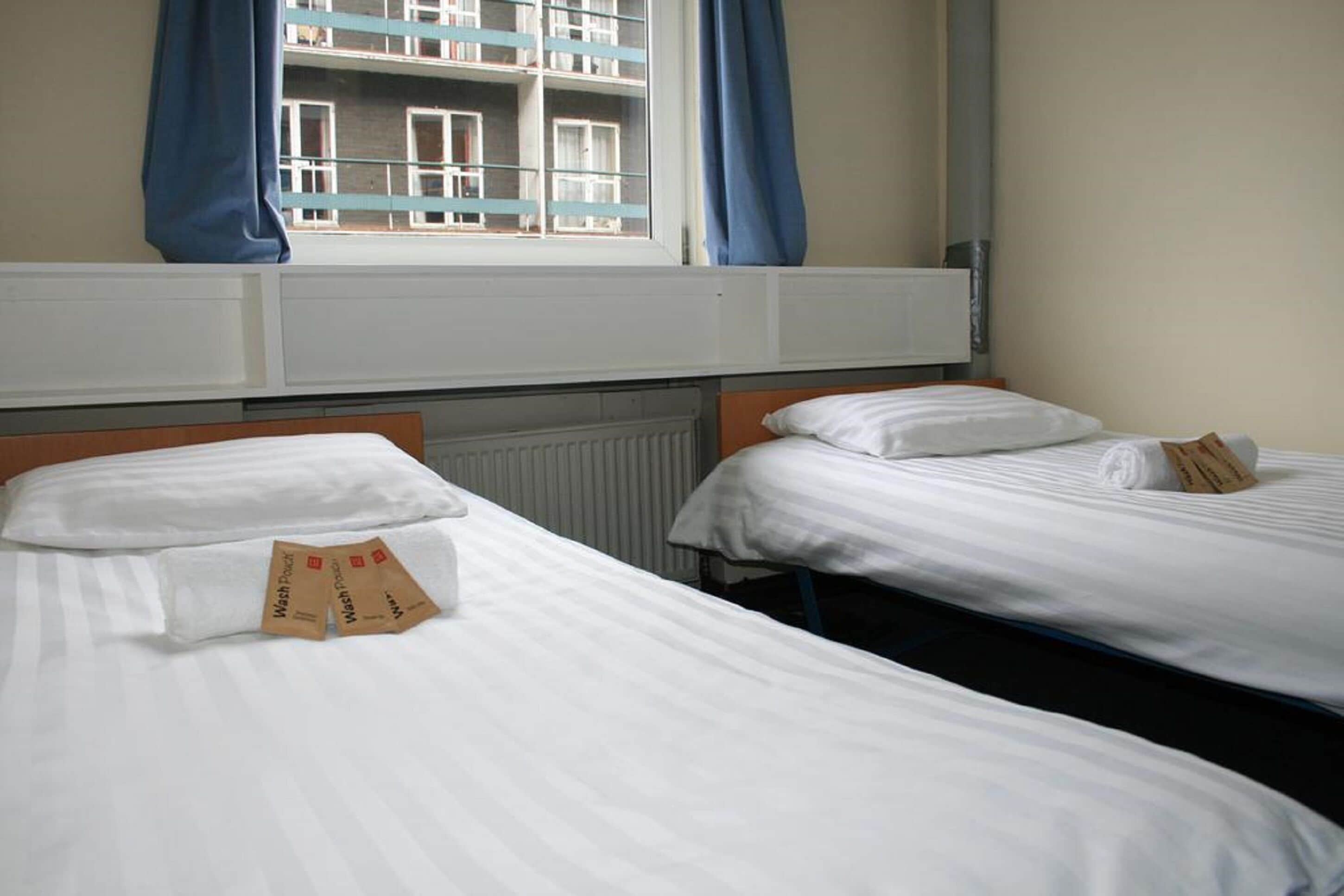 A hotel room with two single beds, white bedding, and a window with blue curtains.