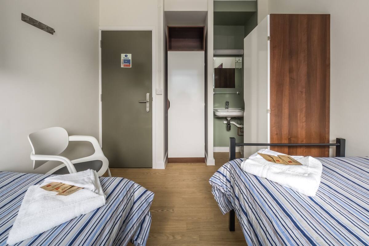 Una habitación de hotel con dos camas, un armario de madera, un baño con espejo y una silla blanca.
