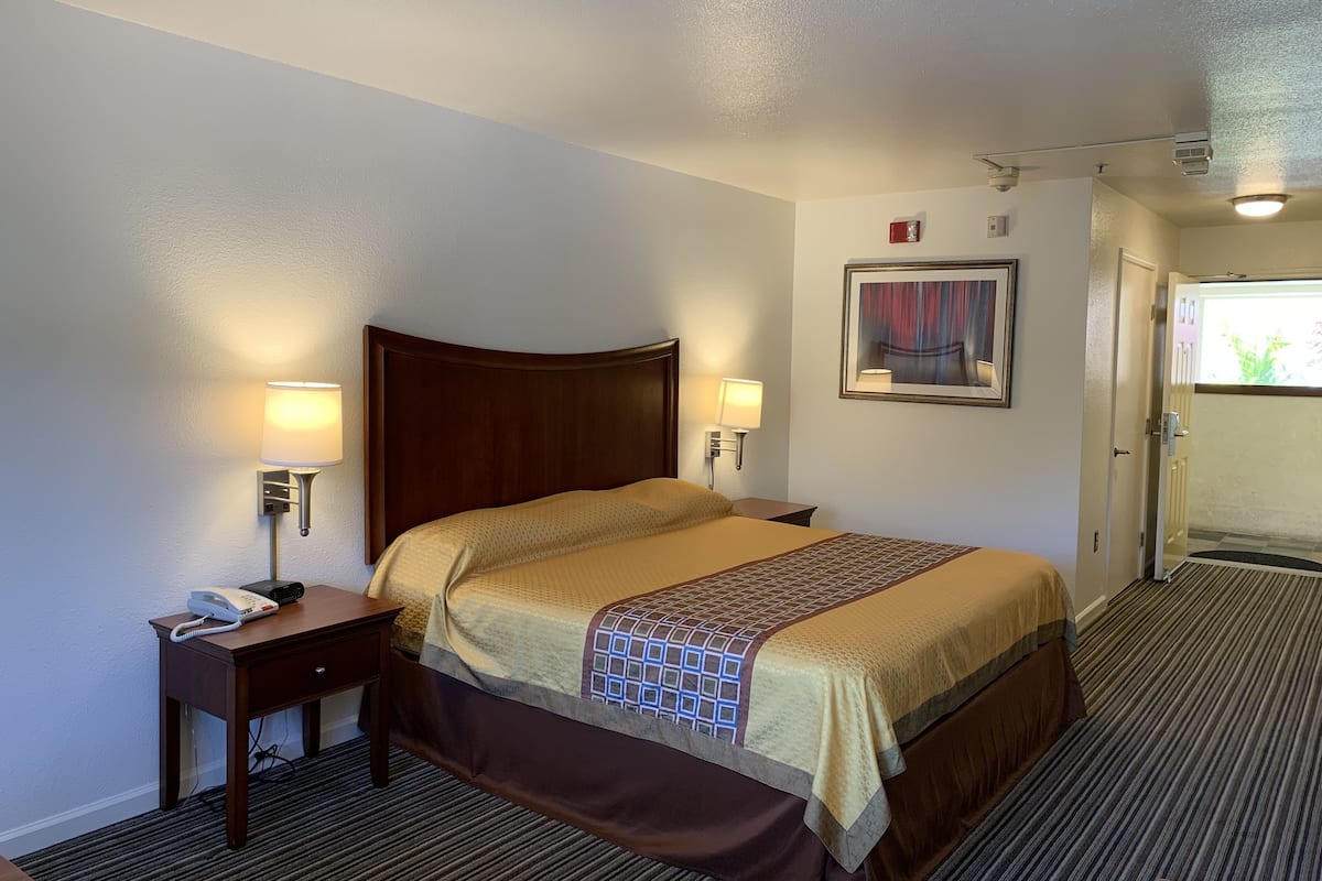 A hotel room with a bed, bedside lamps, a telephone, a painting on the wall, and a door leading to the bathroom.