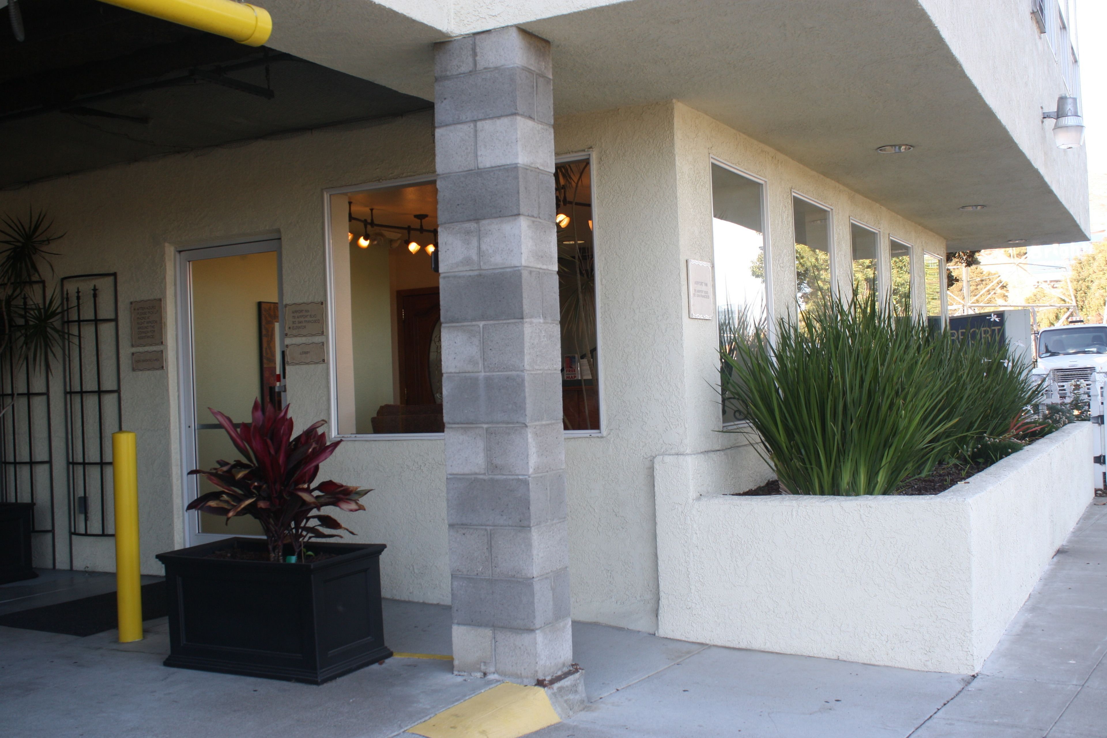 A building with a stone pillar, a planter with plants, and a yellow bollard.