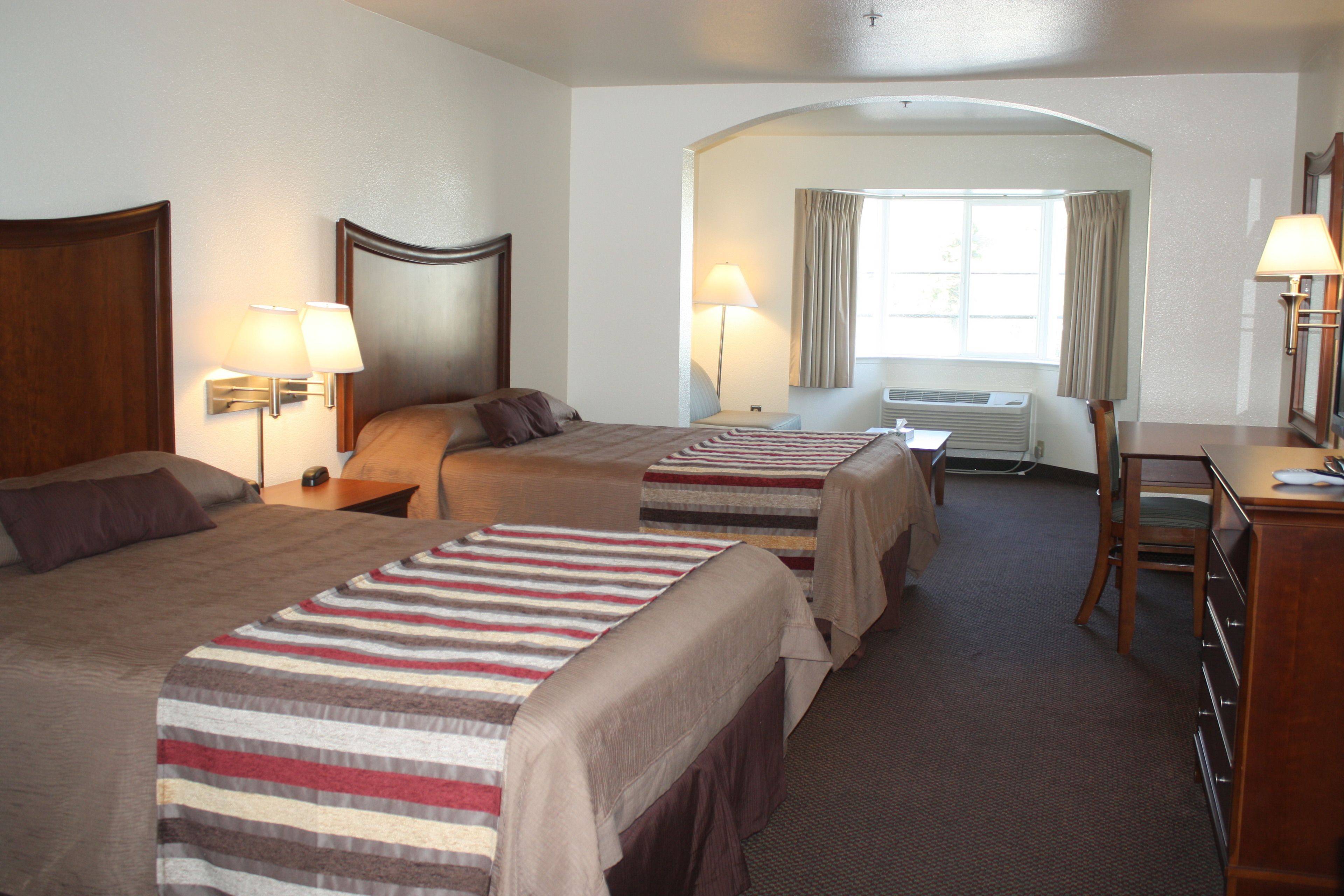 A hotel room with two beds, a desk, a chair, a lamp, and an air conditioning unit.