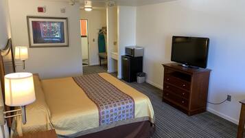 A hotel room with a bed, a television on a wooden stand, a desk with a lamp, a microwave, and a trash bin.