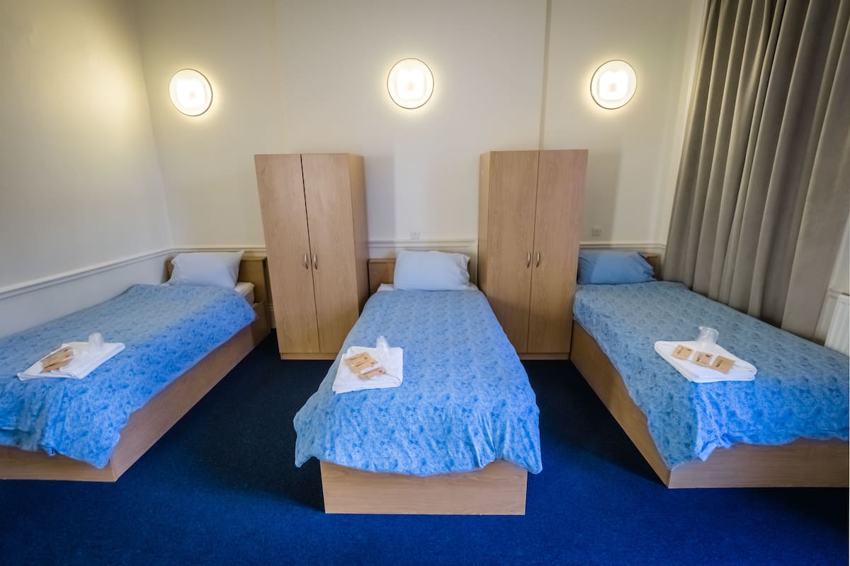 Quarto de hotel com três camas de solteiro, cada uma com uma colcha azul e uma toalha branca. Há armários de madeira e uma luminária redonda na parede sobre cada cama.