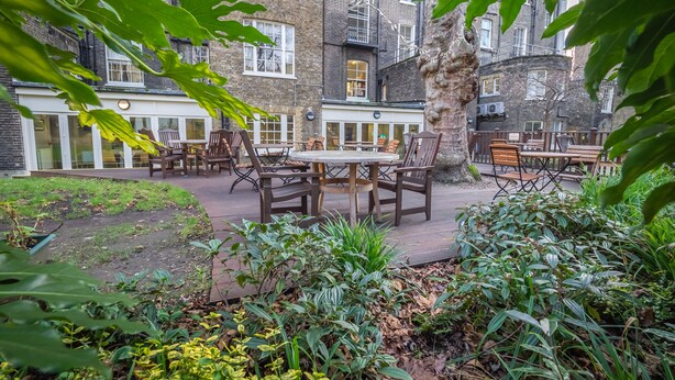 Un espace extérieur aménagé avec des tables et des chaises, entouré de verdure et avec un bâtiment en briques en arrière-plan.