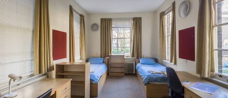 A dormitory room with two single beds, a desk, a chair, and a radiator.