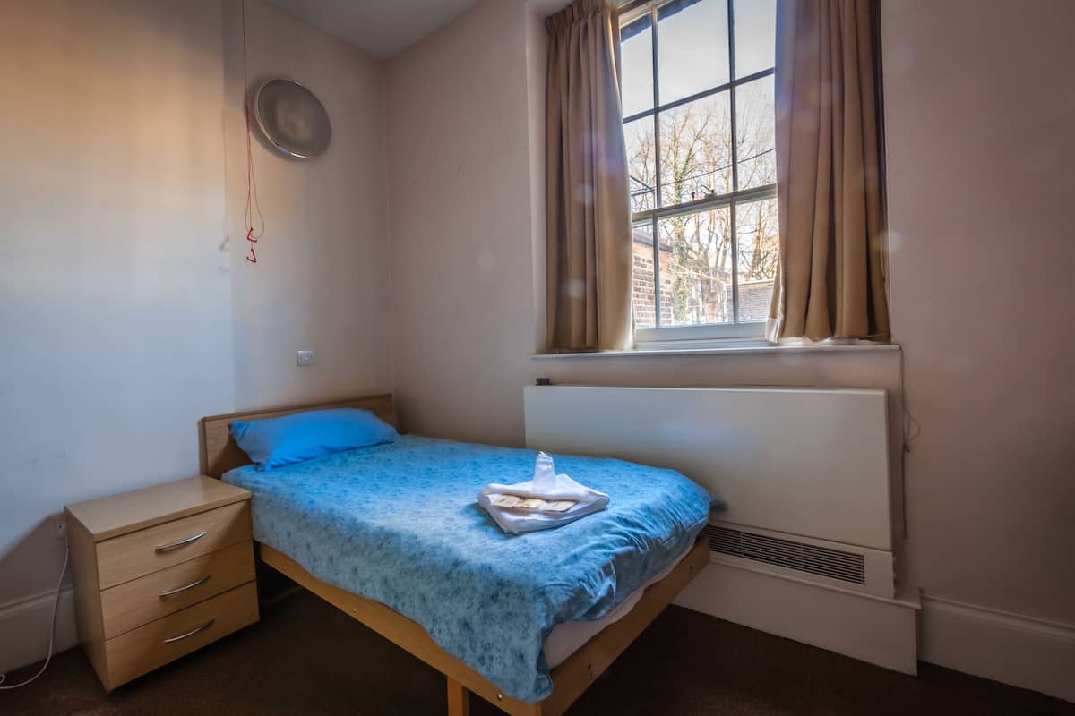 Quarto pequeno com uma cama de solteiro, janela, radiador e mesa de cabeceira.