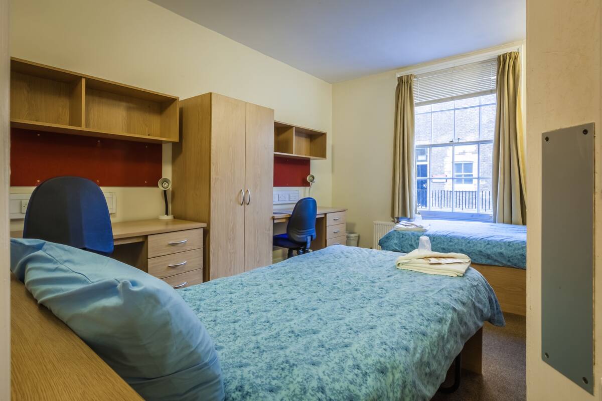 Quarto com duas camas de solteiro, escrivaninha com cadeira, guarda-roupa e janela com cortinas.