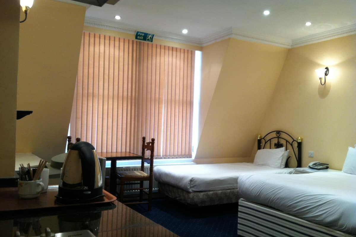 A hotel room with a bed, a chair, a desk with a kettle, and a window with blinds.