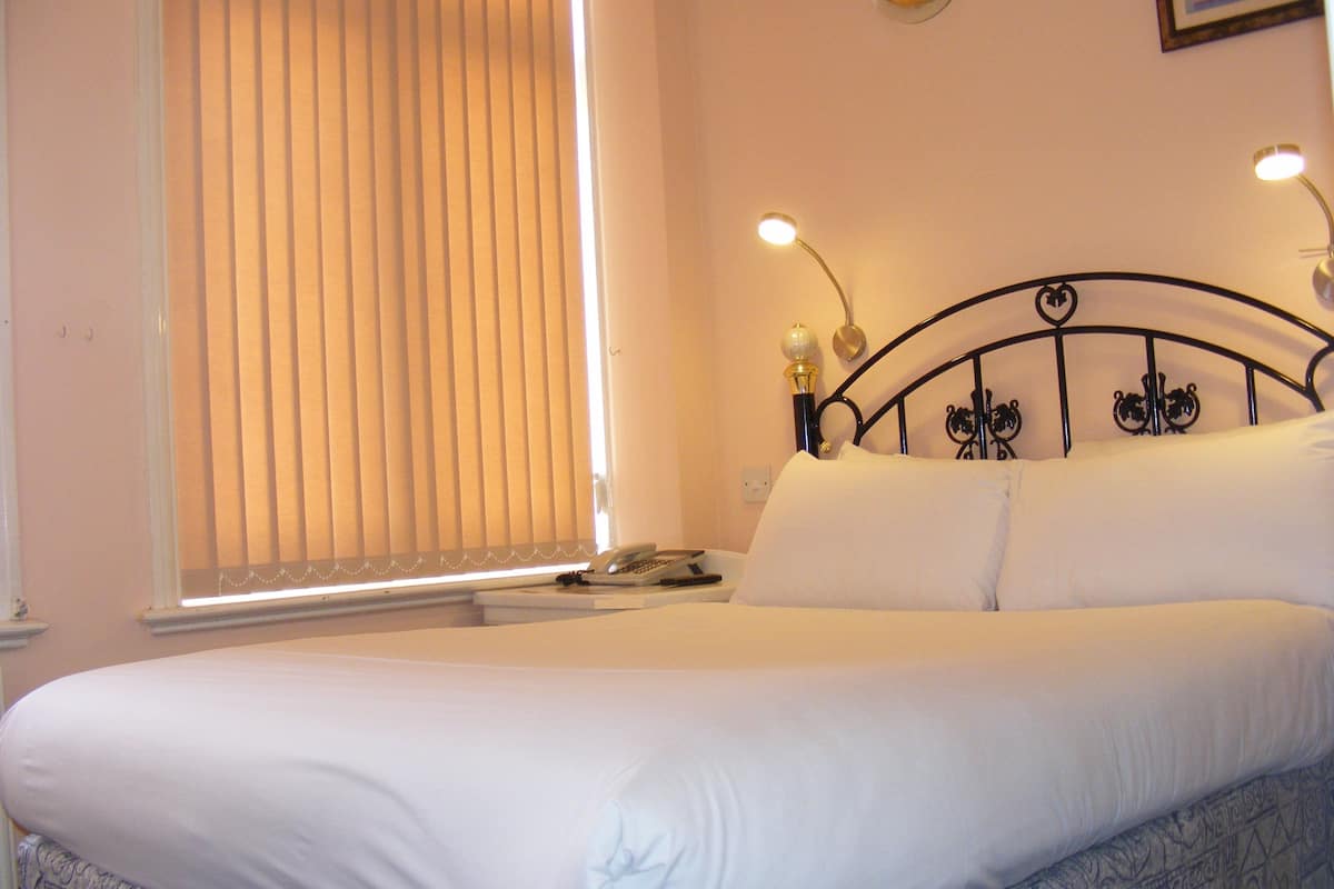 A neatly made bed with white linens, a bedside table with a lamp, and a window with blinds.