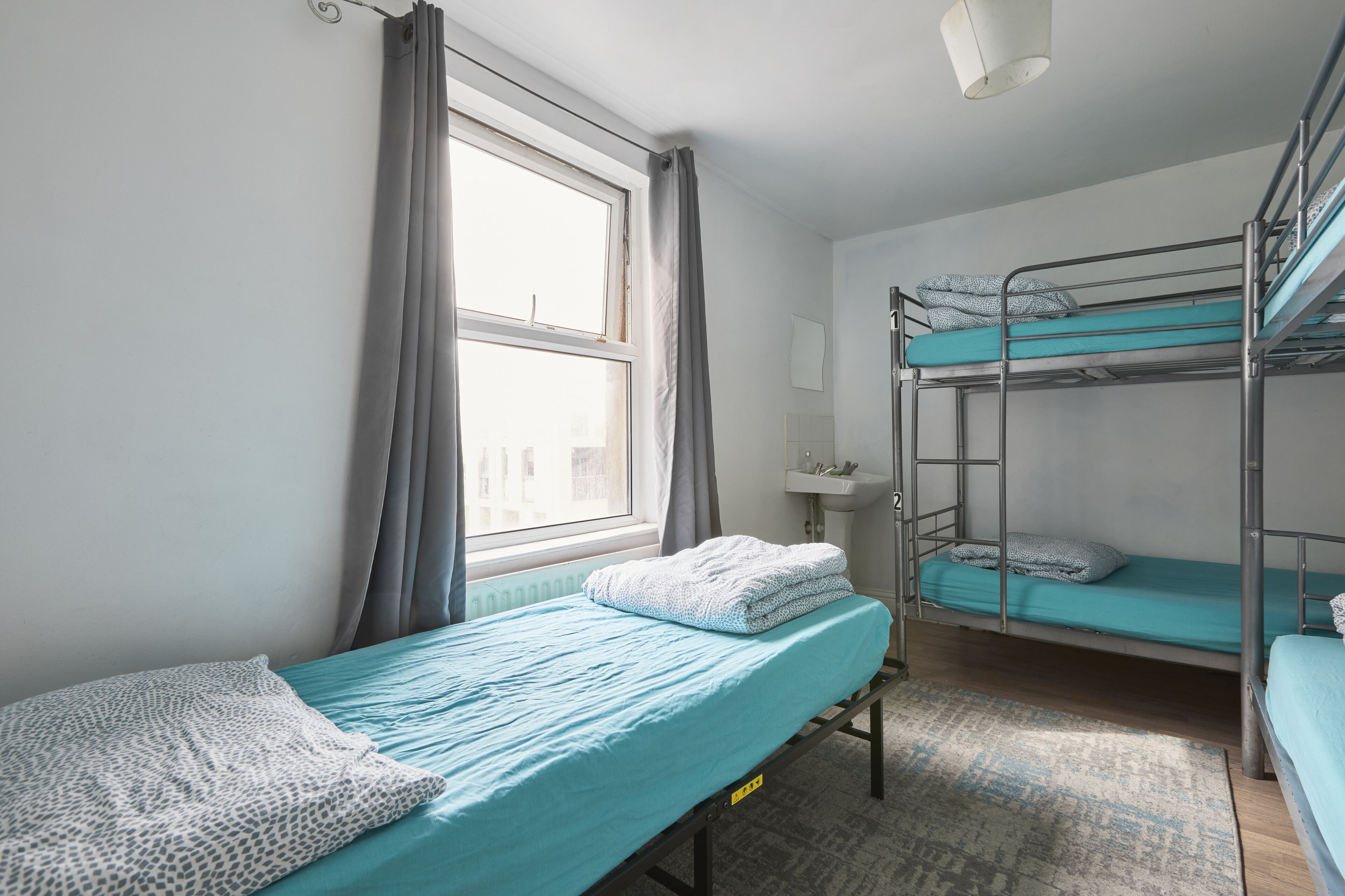 Een slaapzaal met stapelbedden, een raam met gordijnen, een nachtkastje en een klein wastafeltje.