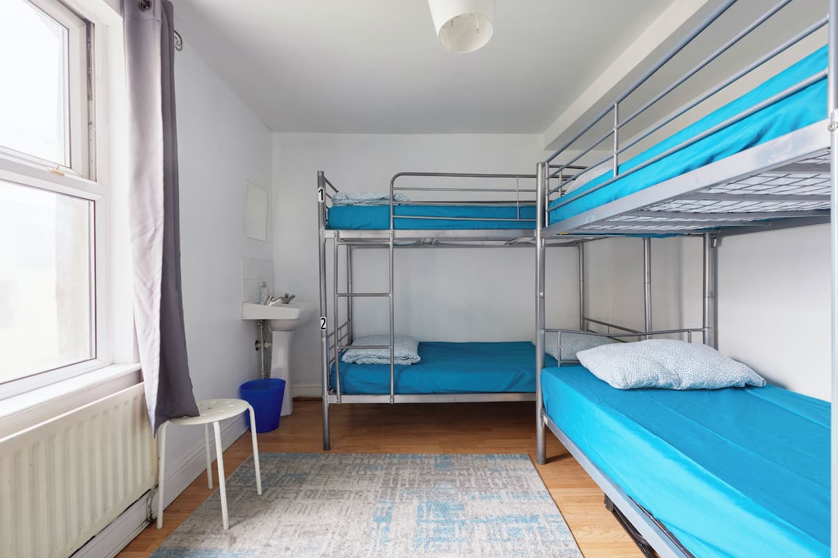 A dormitory room with bunk beds, a small white chair, a sink, and a window with curtains.