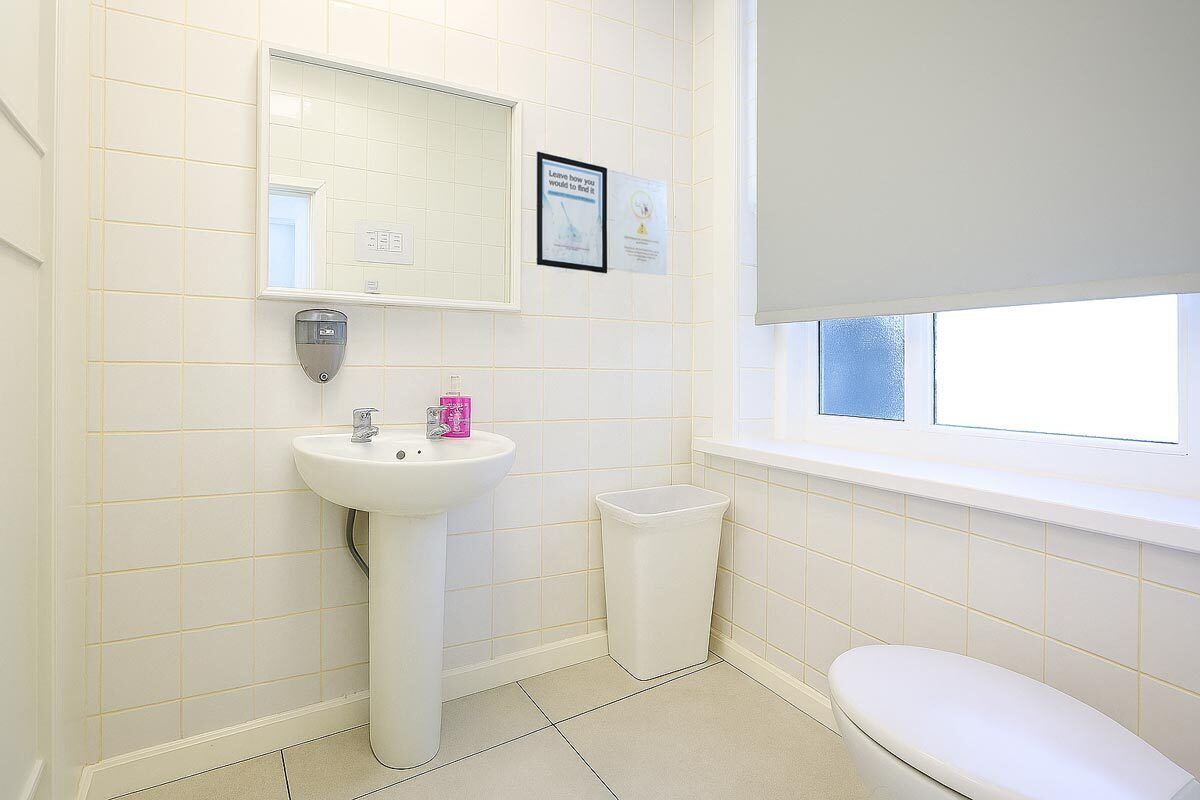 A bathroom with a white sink, mirror, and toilet, a window with a white blind, and a trash bin.