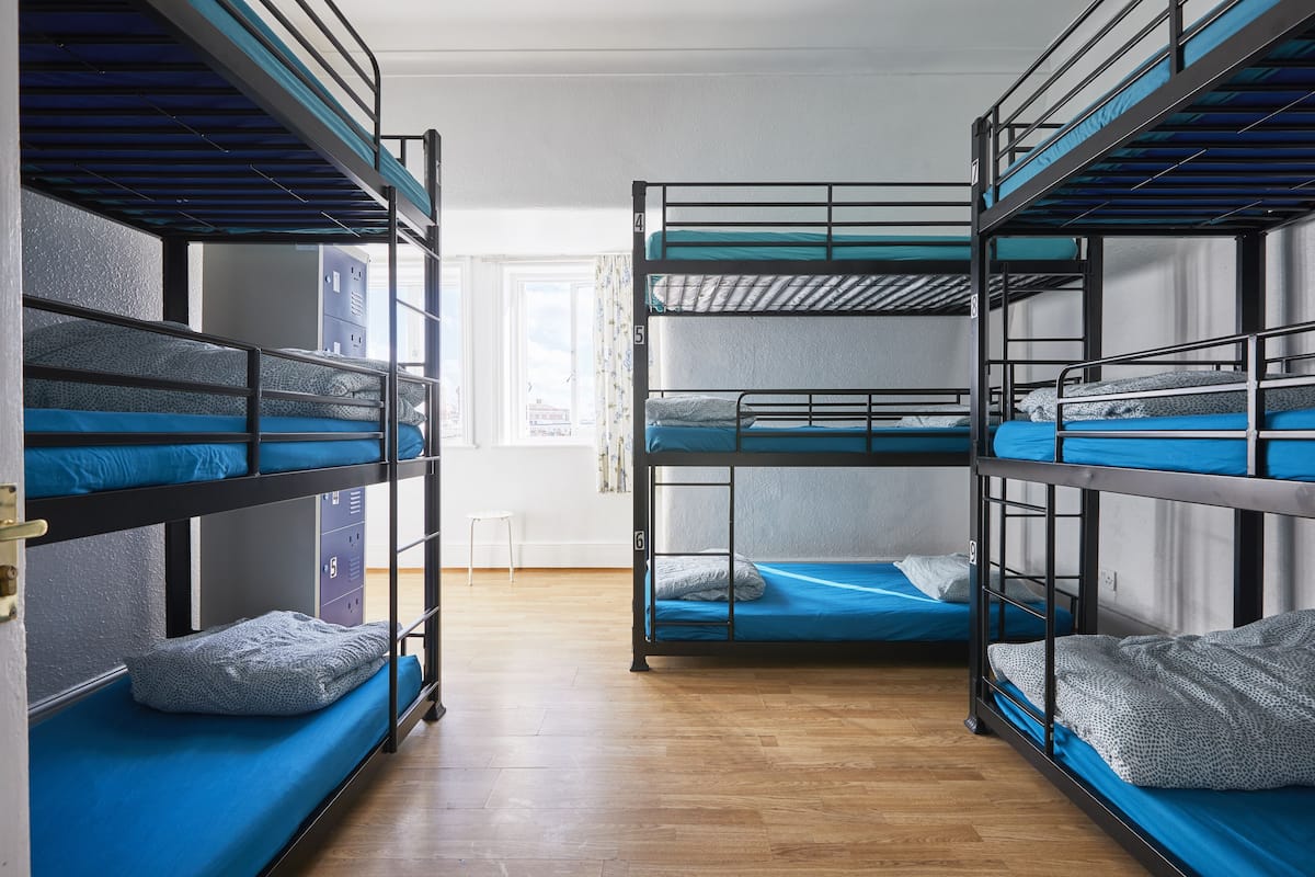 A dormitory room with bunk beds, wooden flooring, and a window with curtains.