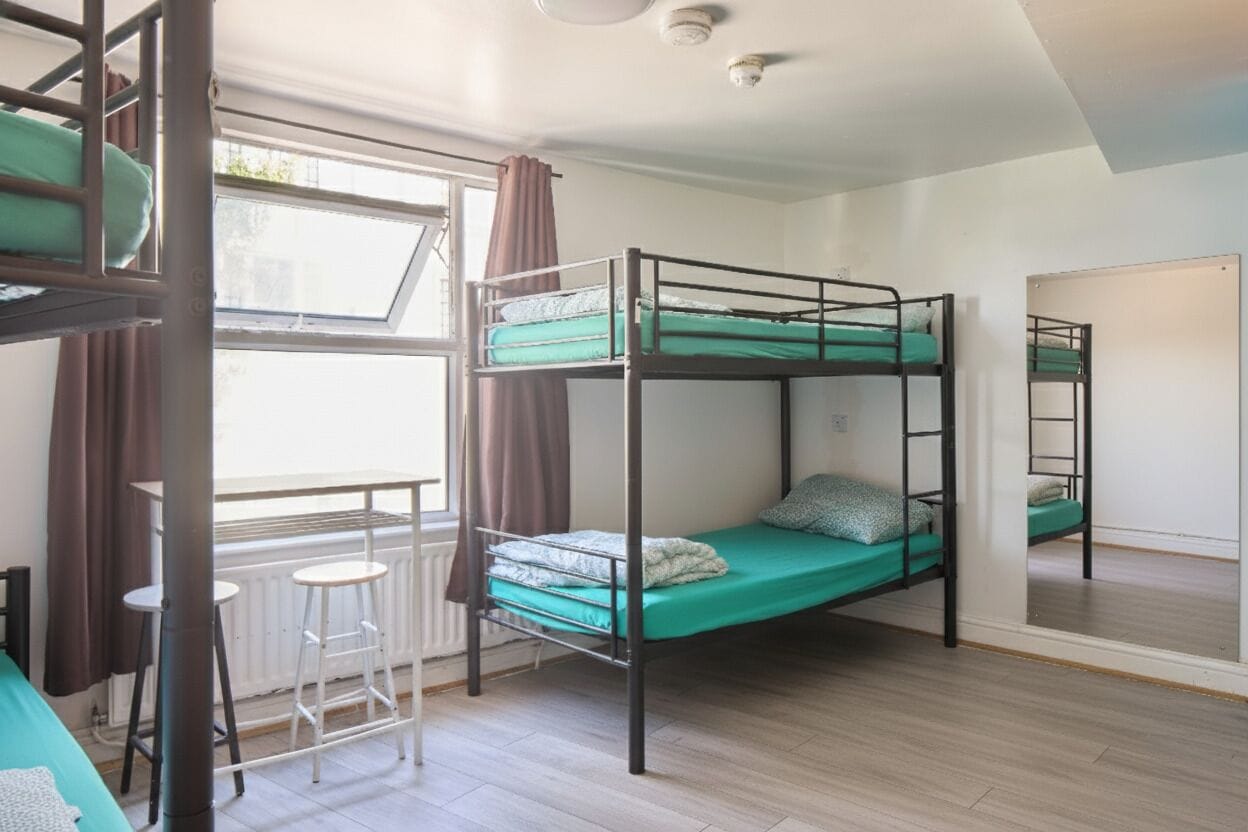 Une chambre de dortoir avec des lits superposés, une fenêtre avec des rideaux, un radiateur et une petite table avec des chaises.