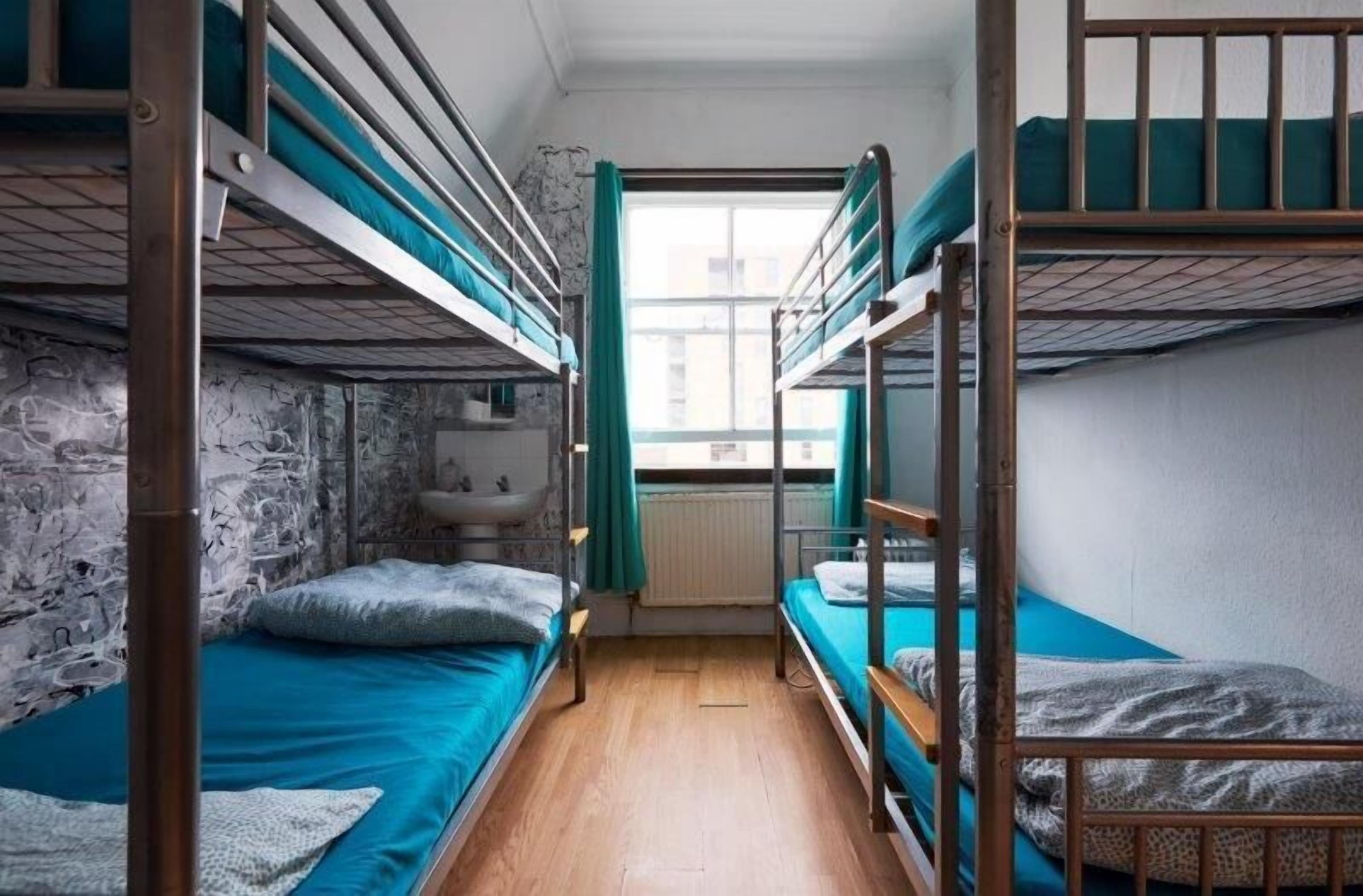 A dormitory room with bunk beds, a window with curtains, a radiator, and a sink.