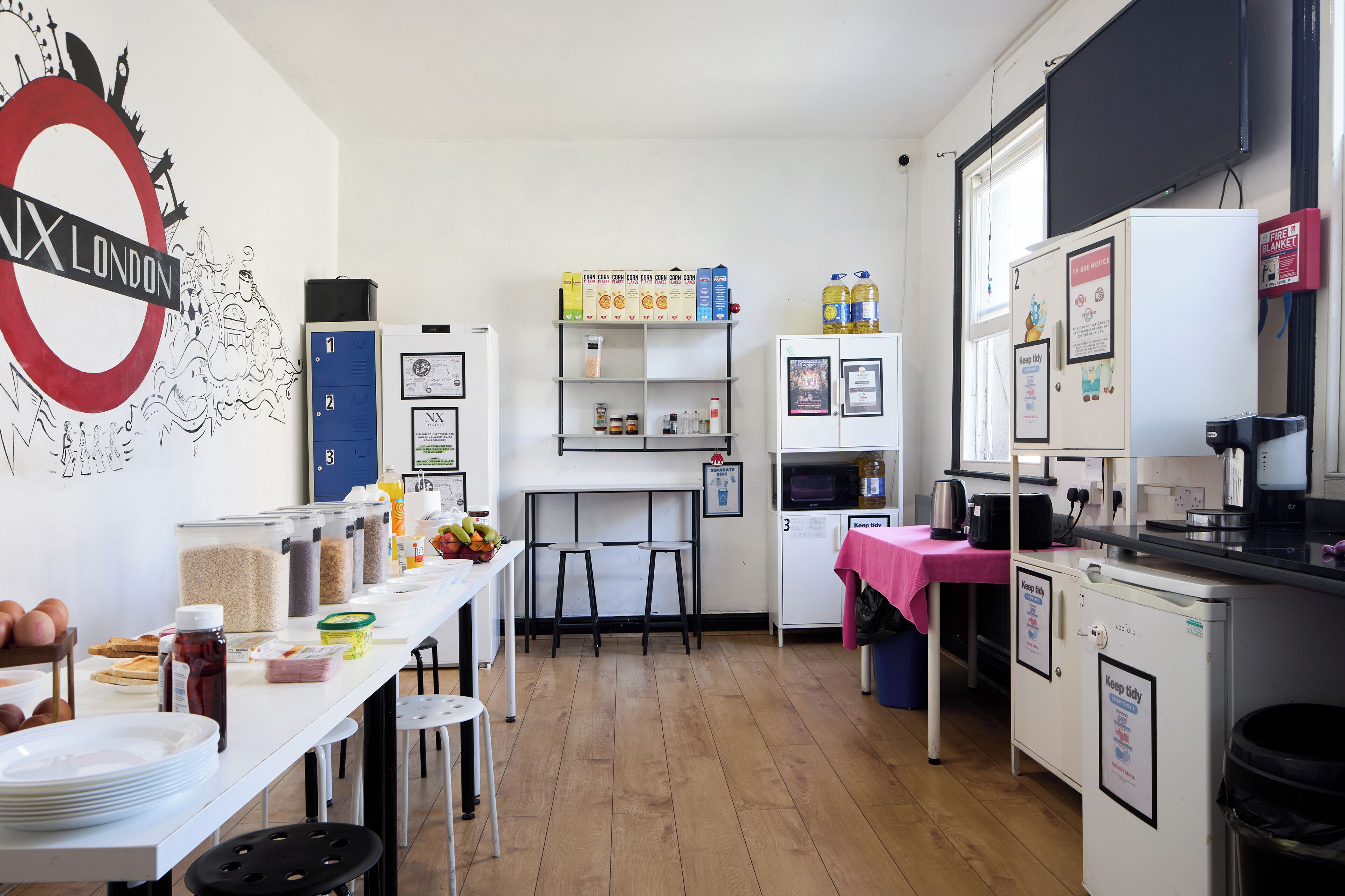A kitchen with a white countertop, a coffee machine, and a refrigerator. There are also shelves with food items and a wall with a London-themed artwork.