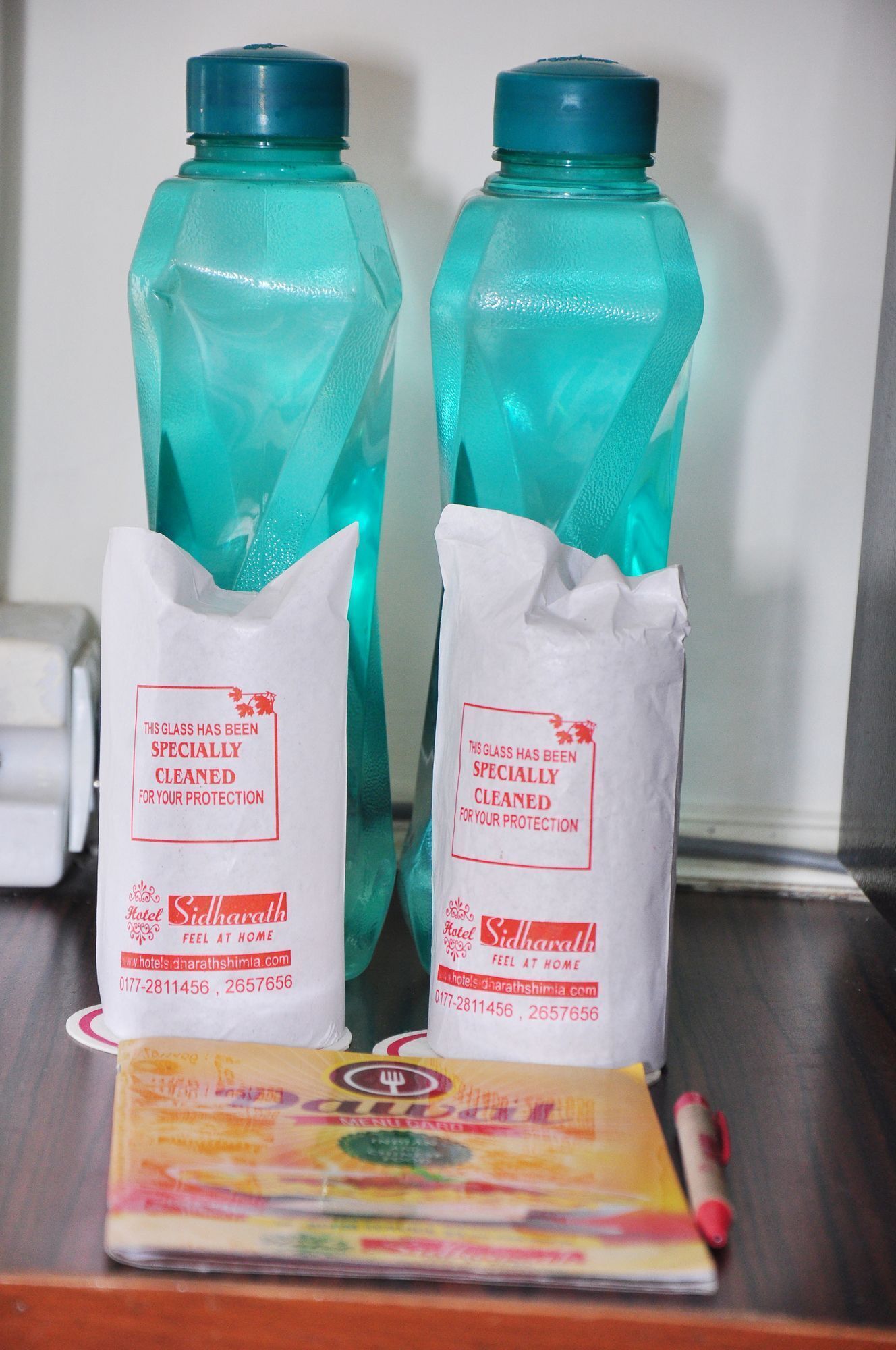 Two turquoise water bottles with white labels, two white bags with red text, and a menu booklet on a wooden surface.