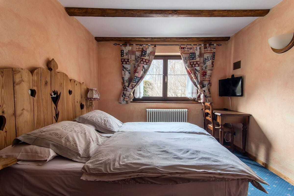 A bedroom with a wooden headboard, a bed with white linens, a window with patterned curtains, a radiator, a desk with a chair, and a television on the wall.