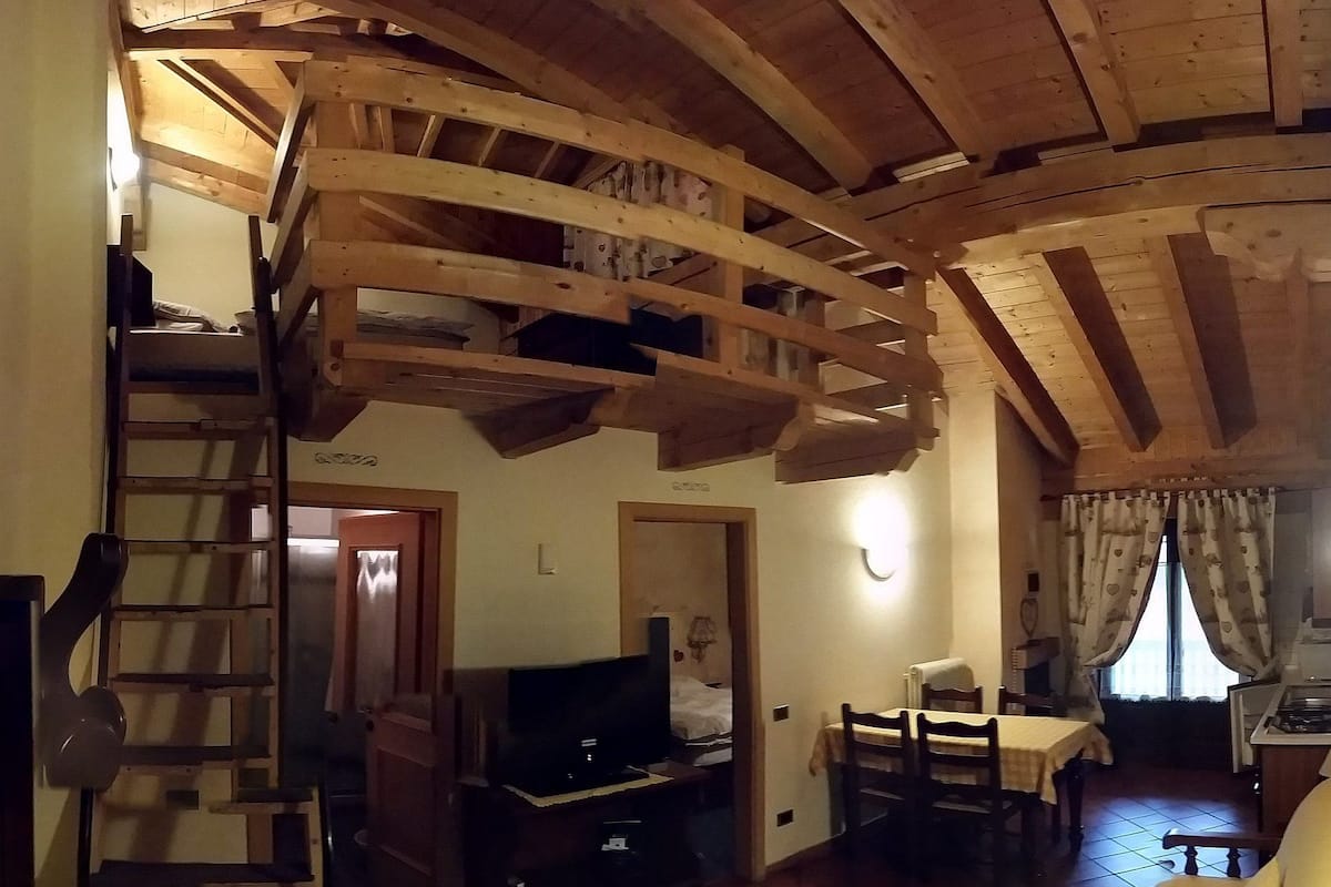 A rustic interior with exposed wooden beams, a staircase, a television, a dining area, and a kitchen.