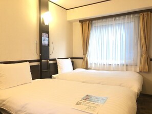 A hotel room with two beds, a window with sheer curtains, a bedside table, and a wall-mounted light.