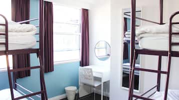 A dormitory room with bunk beds, a desk, and a small window with curtains.