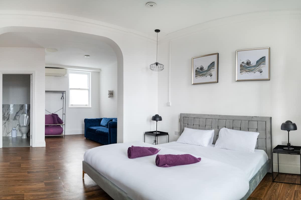 A modern hotel room with a large bed, a blue armchair, a small side table, and a view of the hallway.
