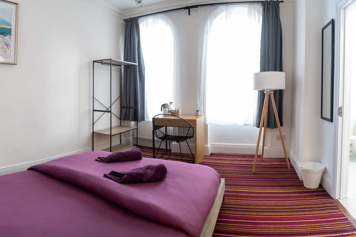 A hotel room with a bed, a desk with a chair, a standing lamp, and a window with curtains.