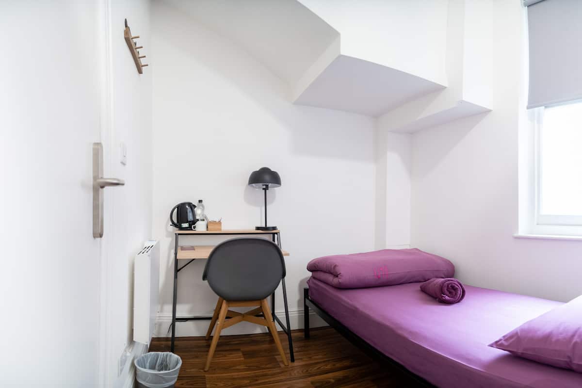 A small, bright room with a desk, chair, and a bed with purple bedding.