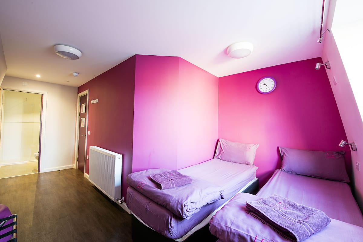 A room with two beds, a radiator, a clock on the wall, and a door leading to a bathroom.