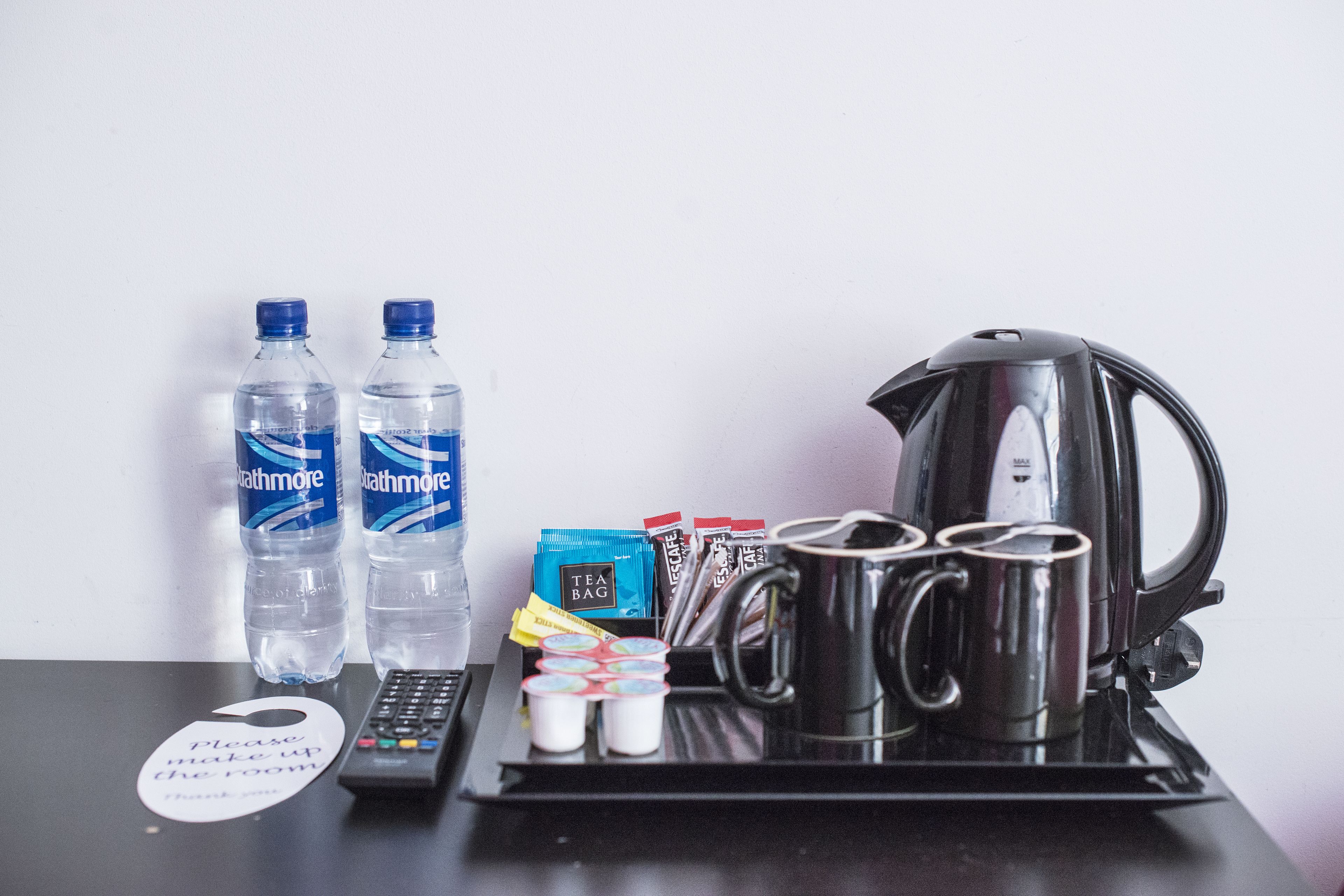 A hotel room's amenities tray with bottled water, tea bags, coffee mugs, a kettle, and a remote control.
