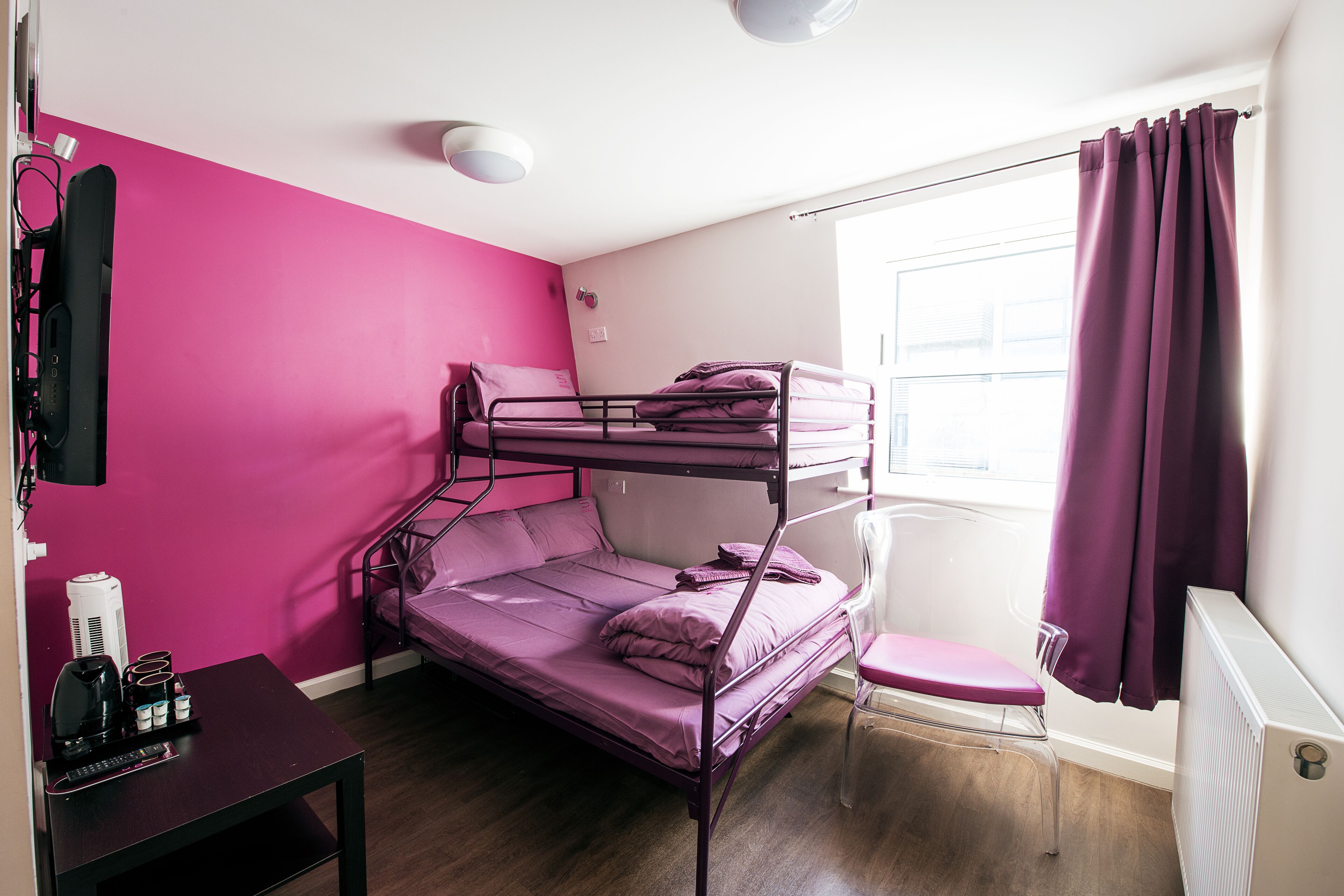 A bunk bed room with pink walls, a window with purple curtains, a radiator, and a desk with a coffee maker.