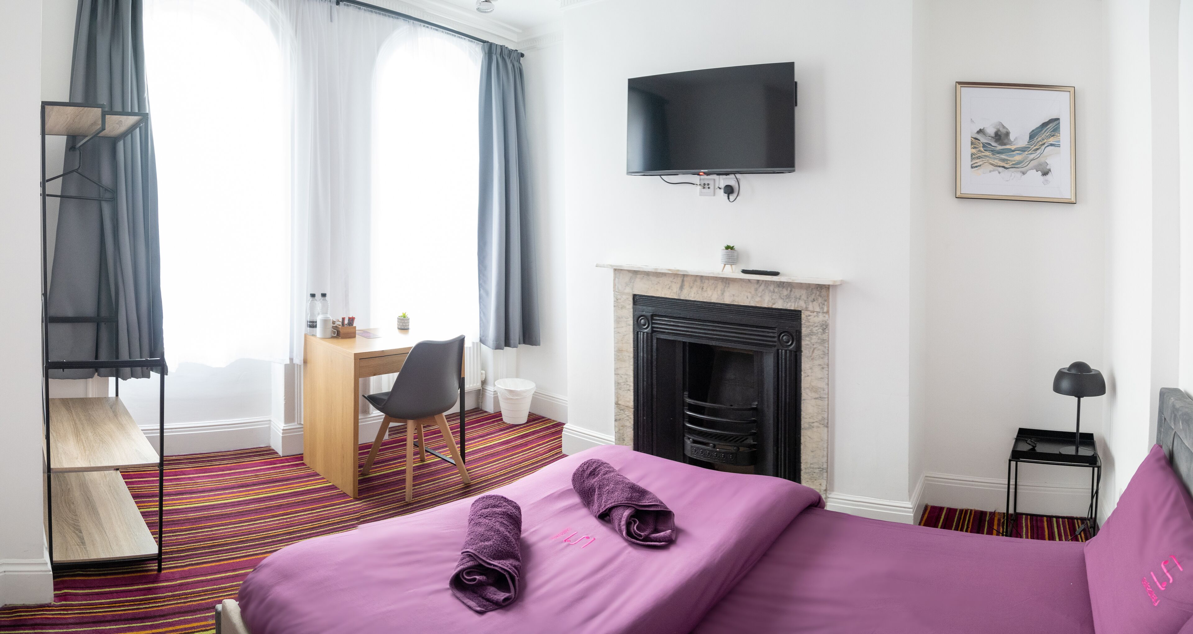 A hotel room with a bed, a desk, a chair, a television, a fireplace, and a painting on the wall.