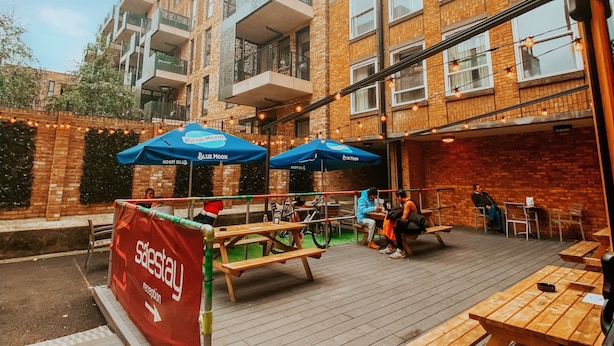 An outdoor patio area with wooden tables and benches, a sign for 'Salestay', and a building with brick walls and balconies in the background. - Safestay London Elephant & Castle (London)