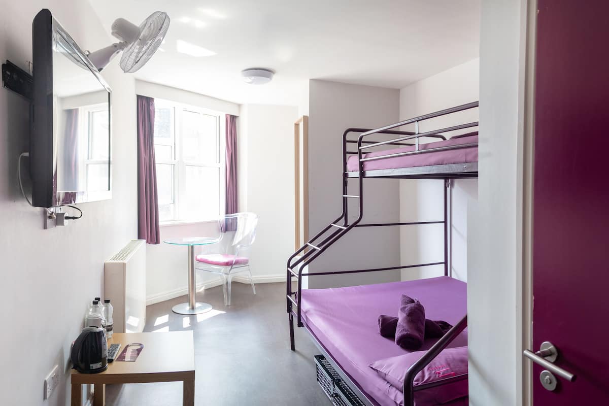 A dormitory room with bunk beds, a desk with a kettle, a chair, and a window with curtains.