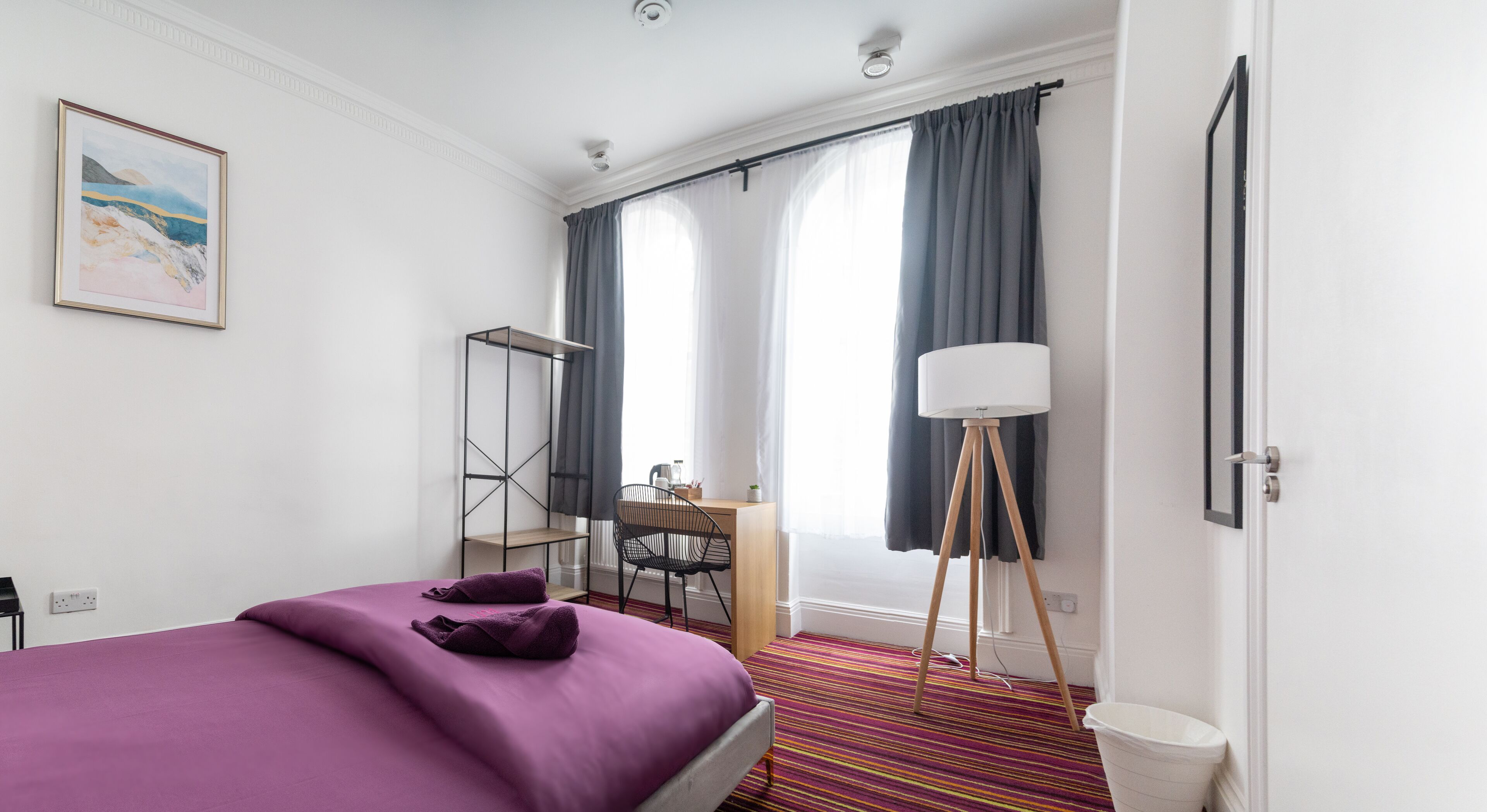 A hotel room with a bed, a desk, a chair, a floor lamp, and a window with curtains.