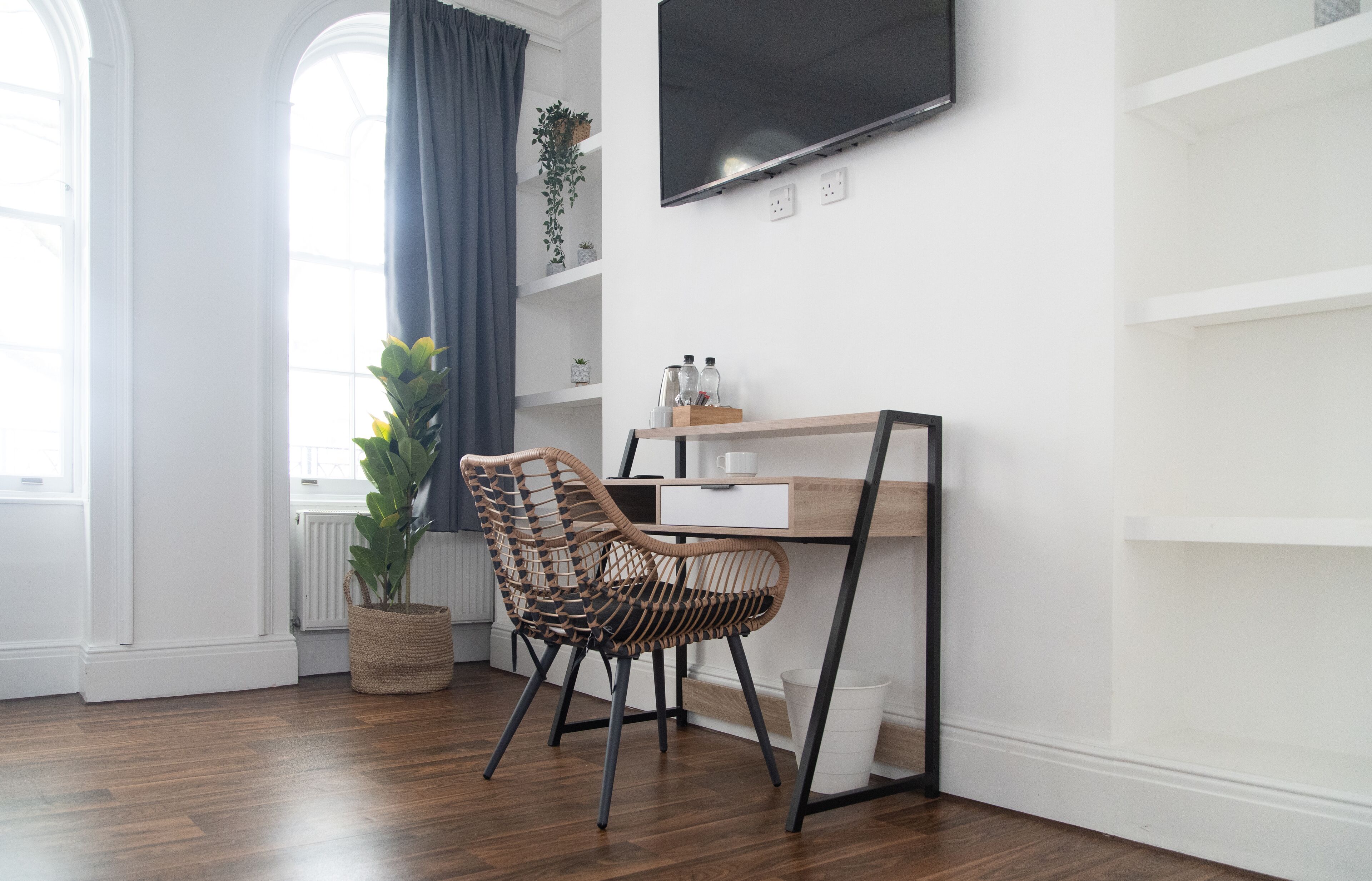 Une chambre moderne avec un bureau en bois, une chaise en osier, une grande fenêtre avec des rideaux, une plante en pot et un téléviseur fixé au mur.